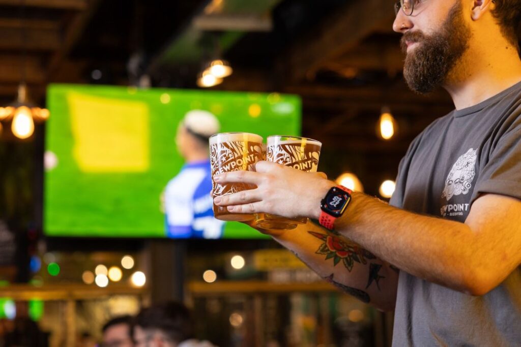 A man carrying 3 beers past a tv showing live sports at a pub
