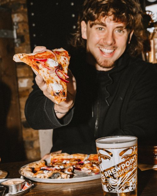 A man holding a pizza slice out in front. There's a beer on the table.